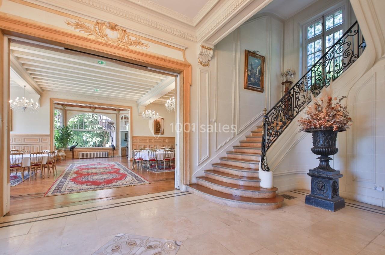 Escalier en bois avec rampe en fer forgé, ouvrant sur une salle lumineuse décorée de lustres et tapis colorés.