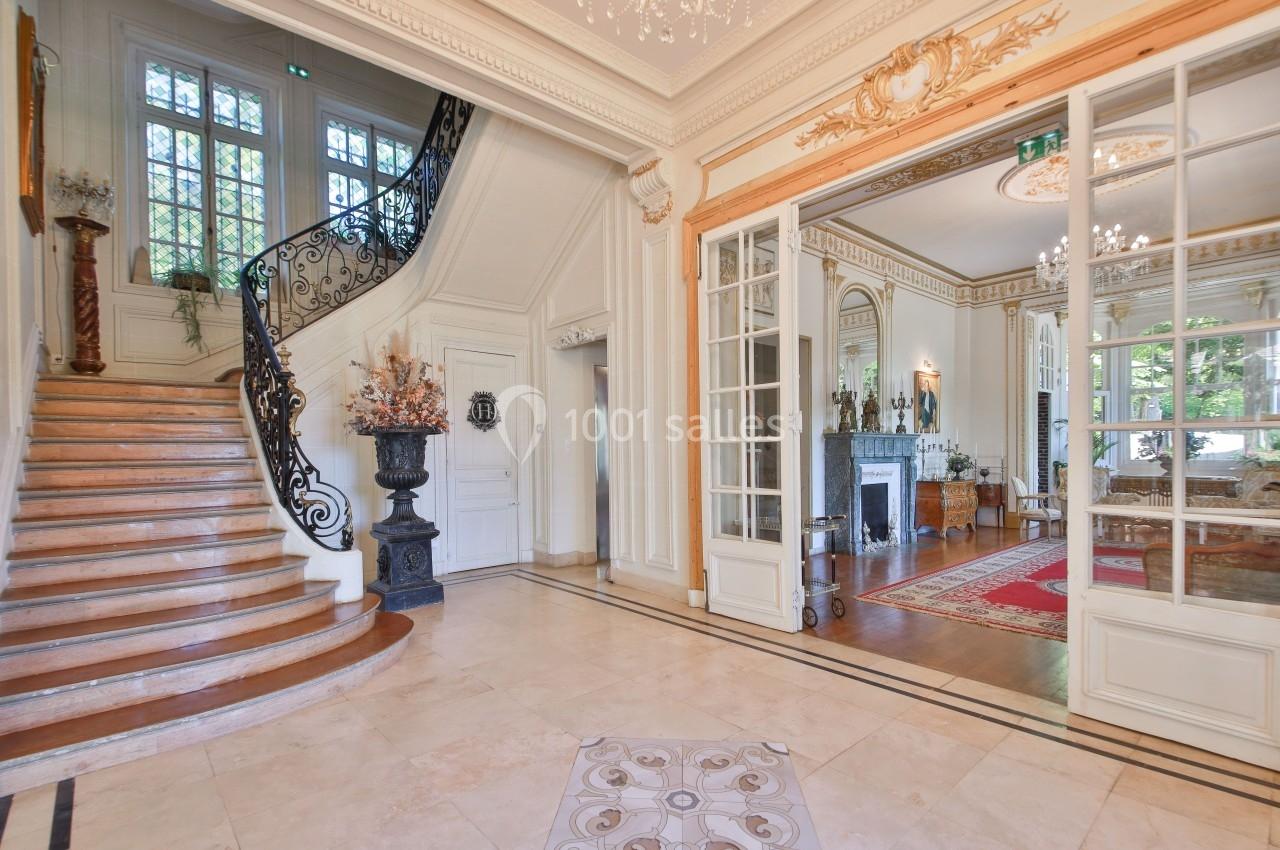 Hall d'entrée lumineux avec escalier en bois, rampe en fer forgé, et vue sur un salon élégant avec cheminée.