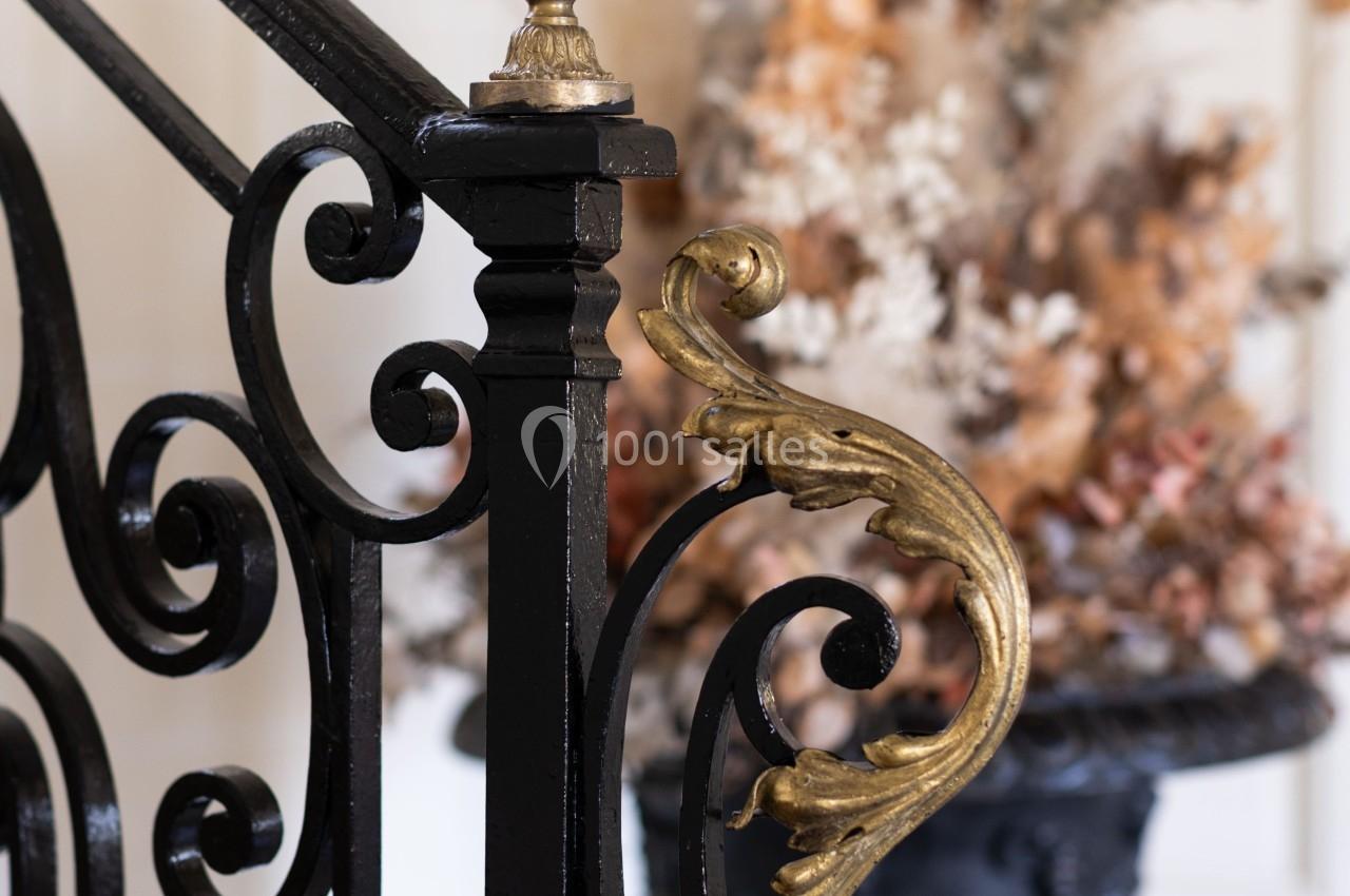 Détail d'une rampe d'escalier en fer forgé noir avec ornements dorés, vase décoratif en arrière-plan flou.