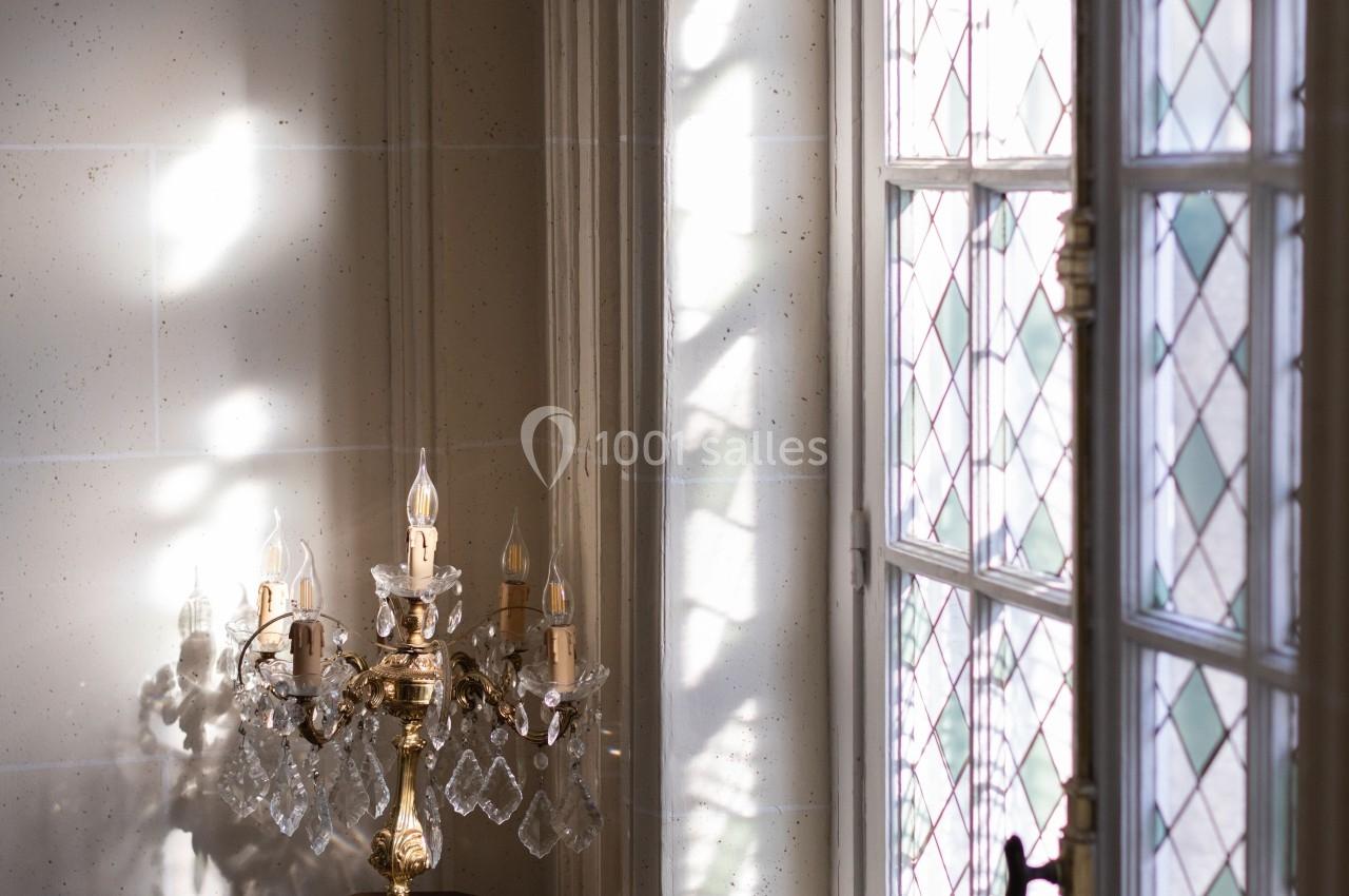 Lumière naturelle traversant une fenêtre à vitraux, éclairant un chandelier en cristal sur un pied en marbre.