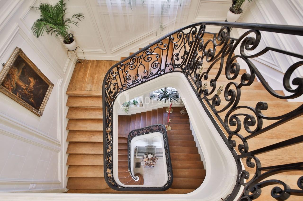 Escalier en bois vu d'en haut avec une rampe en fer forgé ornée, entouré de plantes et d'un tableau mural.