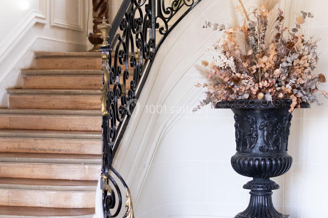 Escalier en bois avec rampe en fer forgé, grand vase décoratif et lumière naturelle entrant par une fenêtre.