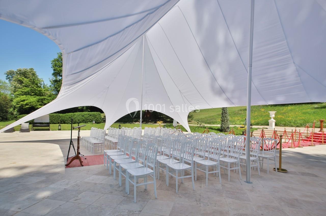 Chaises blanches alignées sous une grande tente blanche dans un espace extérieur pavé, entouré de verdure.