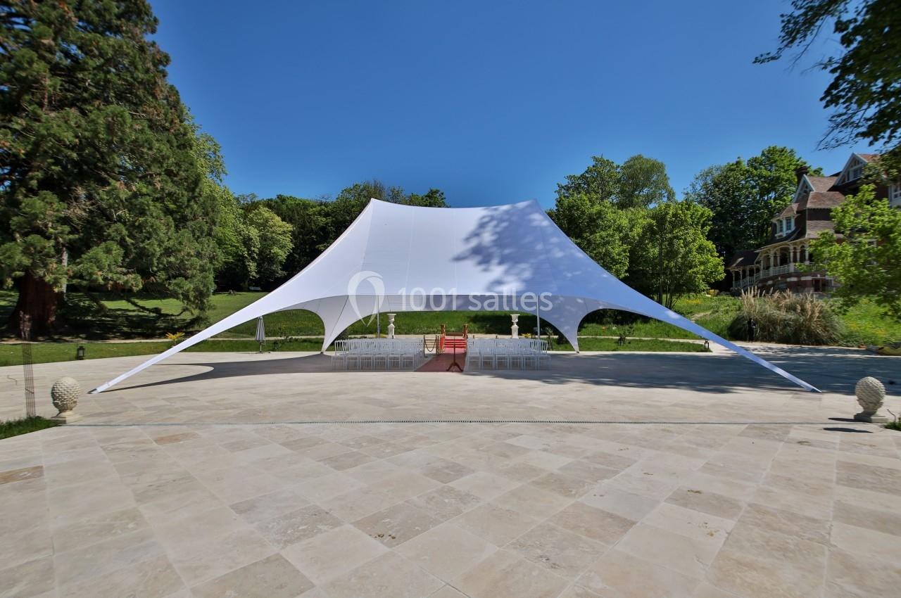 Tente blanche tendue sur une grande terrasse en pierre, entourée de verdure et d'arbres sous un ciel dégagé.