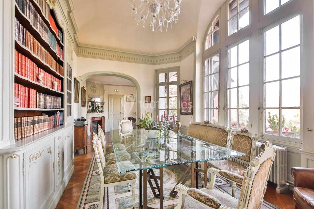 Salle à manger élégante avec table en verre, chaises classiques, bibliothèque garnie et grandes fenêtres laissant entrer la…