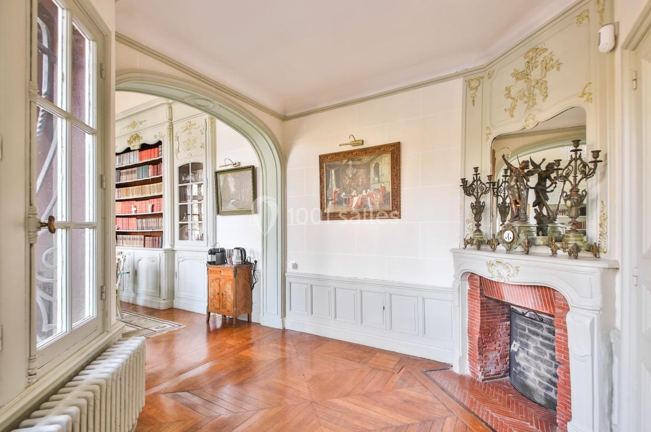Intérieur lumineux avec parquet, cheminée ornée, bibliothèque et moulures décoratives sur les murs.