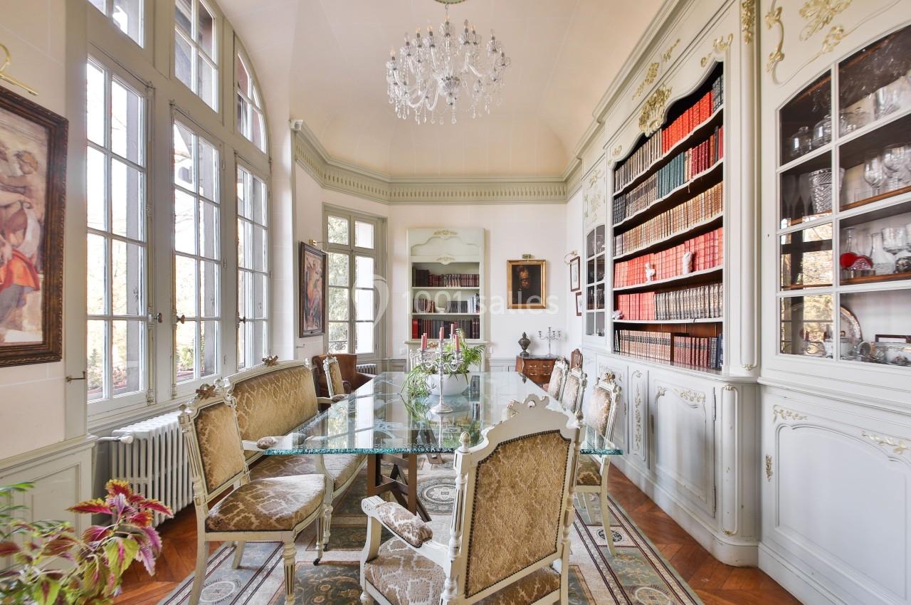 Salle à manger élégante avec table en verre, chaises ornées, bibliothèque remplie de livres et lustre en cristal.