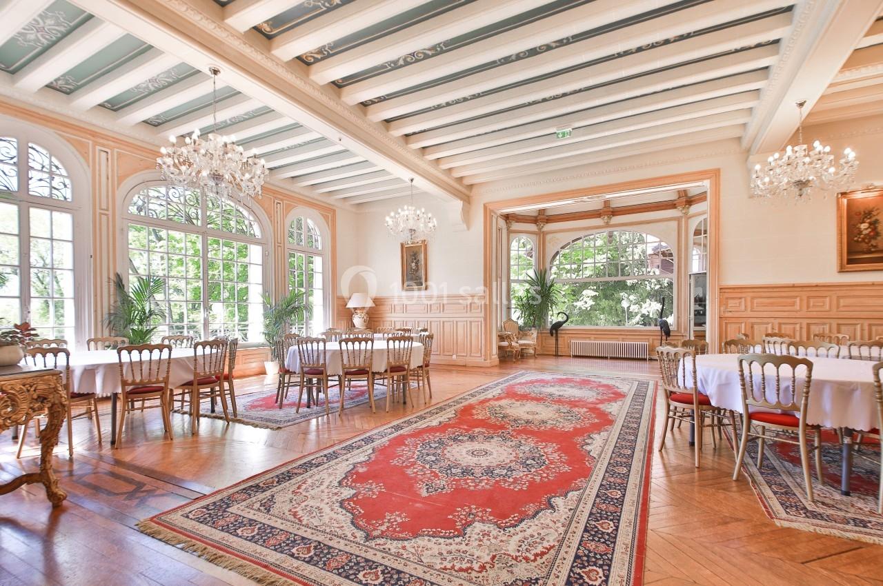 Salle lumineuse avec grandes fenêtres, lustres, tables rondes dressées, tapis rouge et parquet en bois clair.
