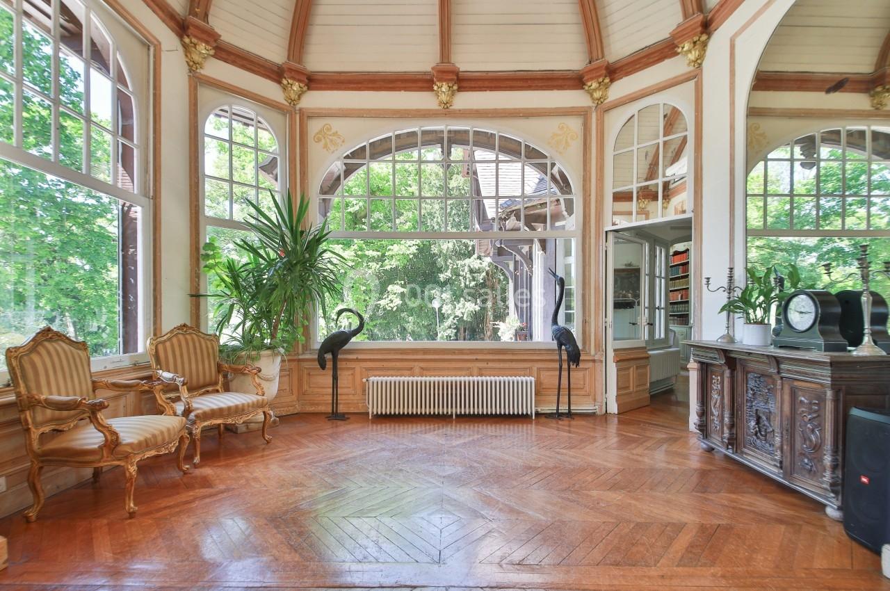 Salle lumineuse avec parquet en bois, grandes fenêtres cintrées, fauteuils anciens et plantes décoratives.