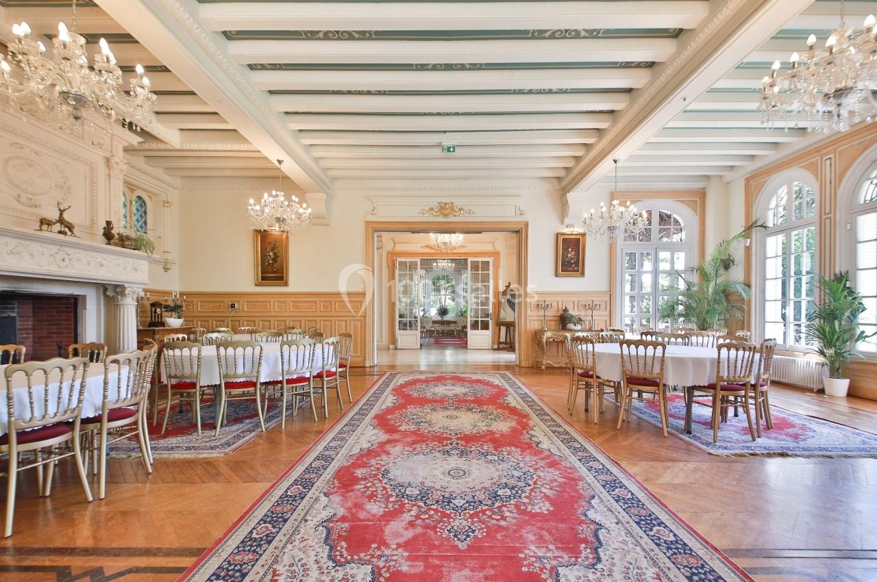 Salle élégante avec tables dressées, tapis rouge central, lustres suspendus et grandes fenêtres laissant entrer la lumière.
