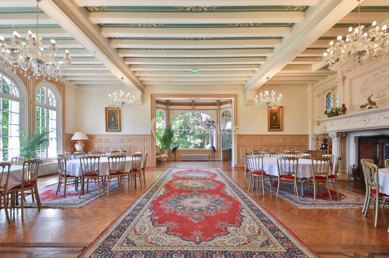 Salle élégante avec tables dressées, lustres, tapis rouge central et cheminée ornée dans un cadre lumineux.
