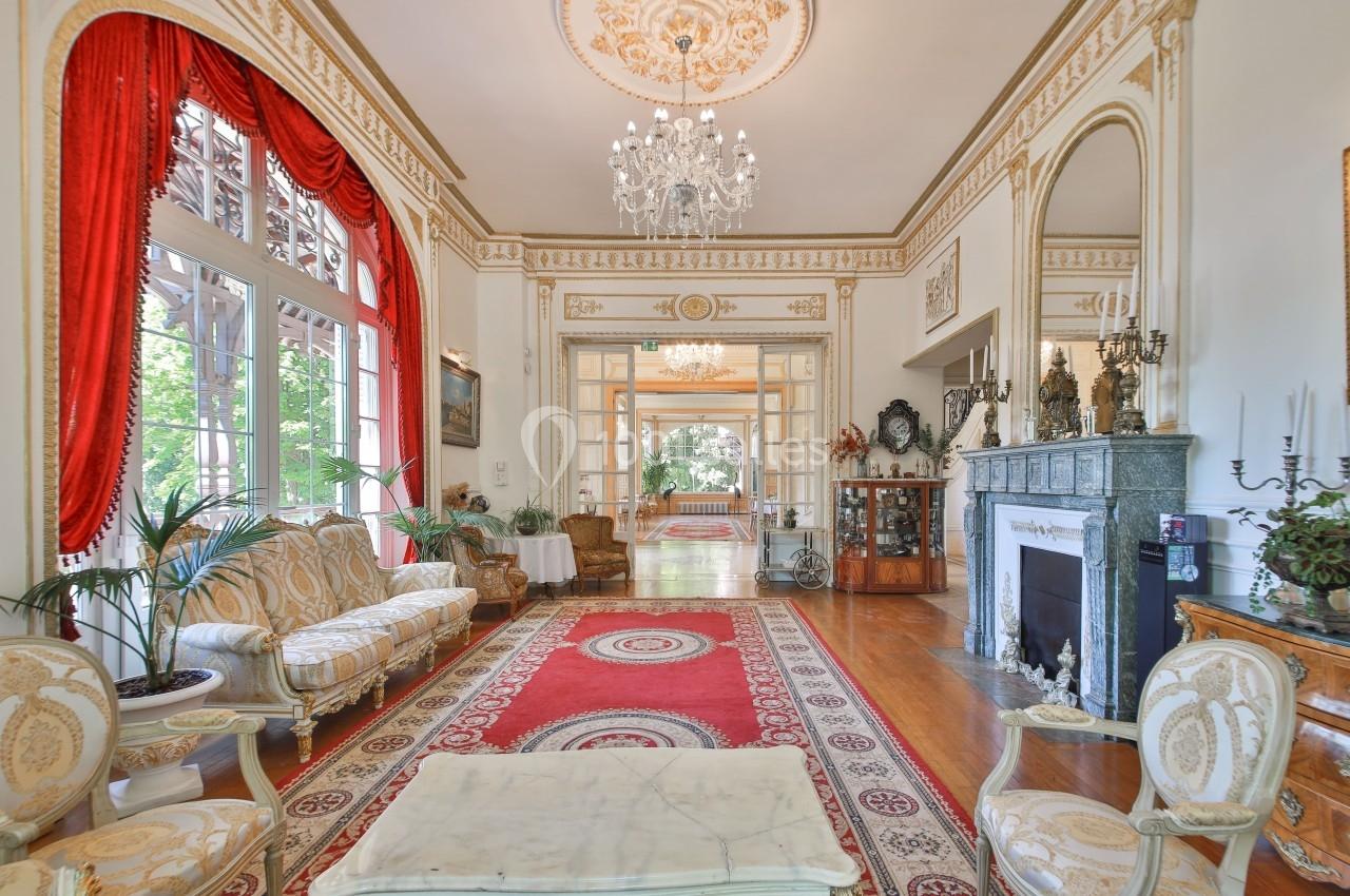 Salon élégant avec mobilier classique, cheminée en marbre, lustres, tapis rouges et grandes fenêtres donnant sur un jardin.