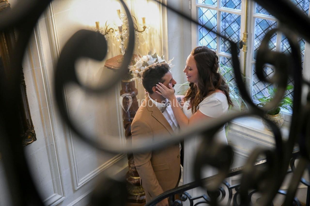Une femme et un homme se regardent tendrement dans un escalier, encadrés par une rambarde en fer forgé.