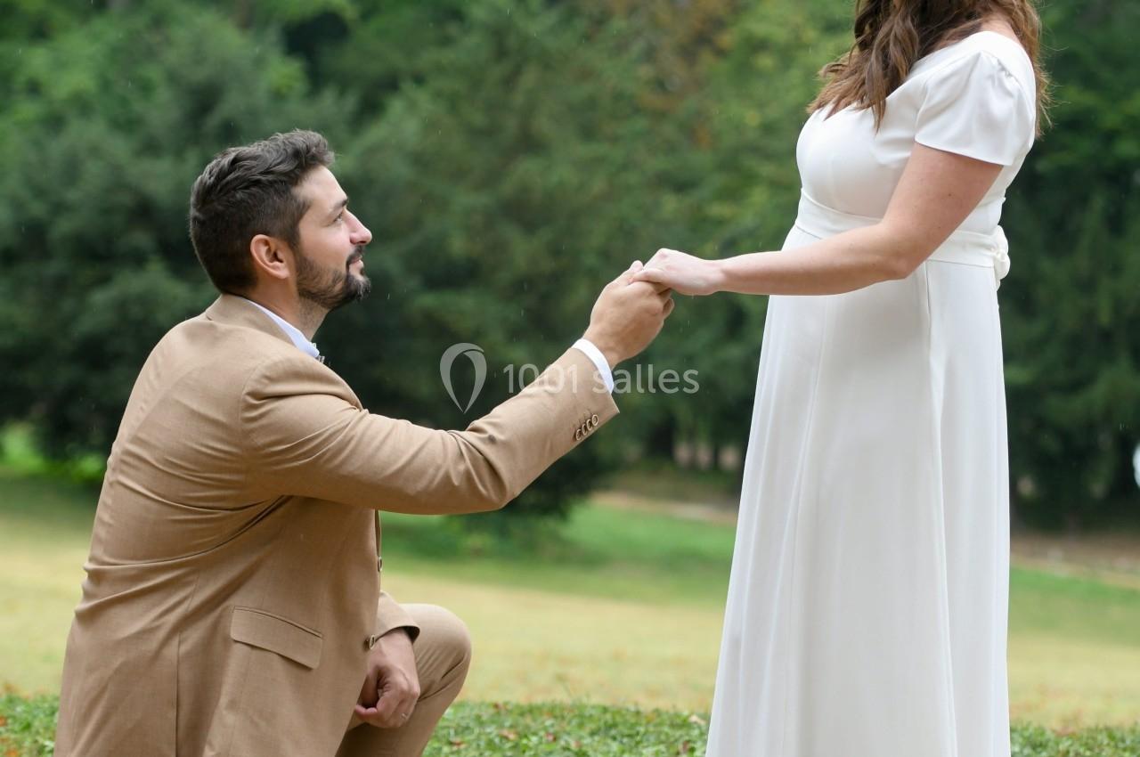 Un homme en costume beige, à genoux, tient la main d'une femme en robe blanche dans un parc verdoyant.