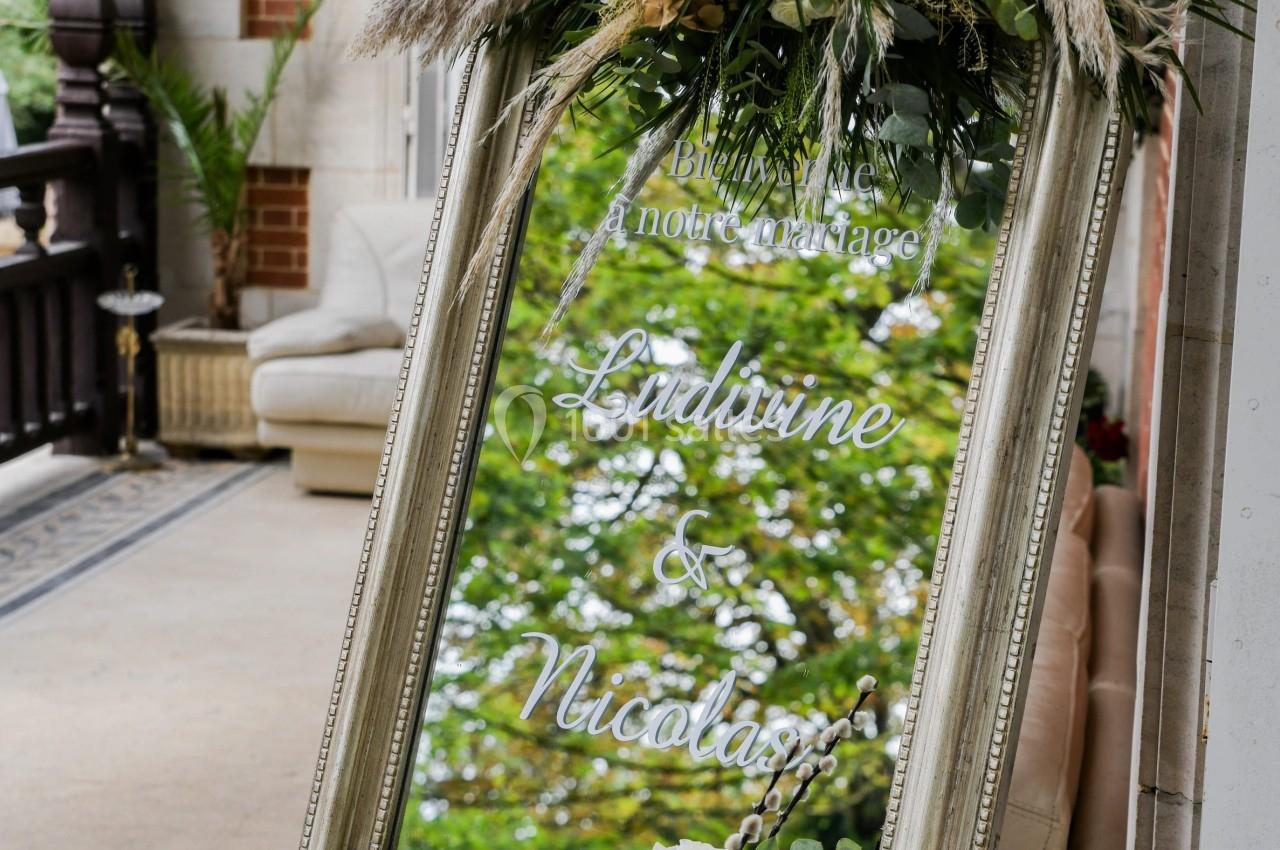 Miroir décoratif avec fleurs et feuillages, affichant des noms gravés, placé sur une terrasse élégante.