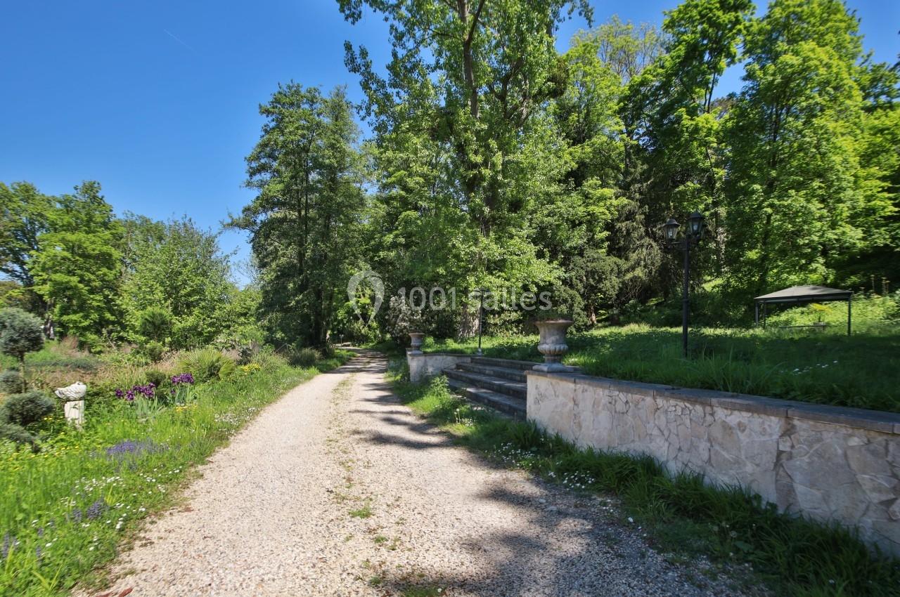 Chemin de gravier bordé de verdure et d'arbres, avec un muret en pierre et des marches sur la droite.