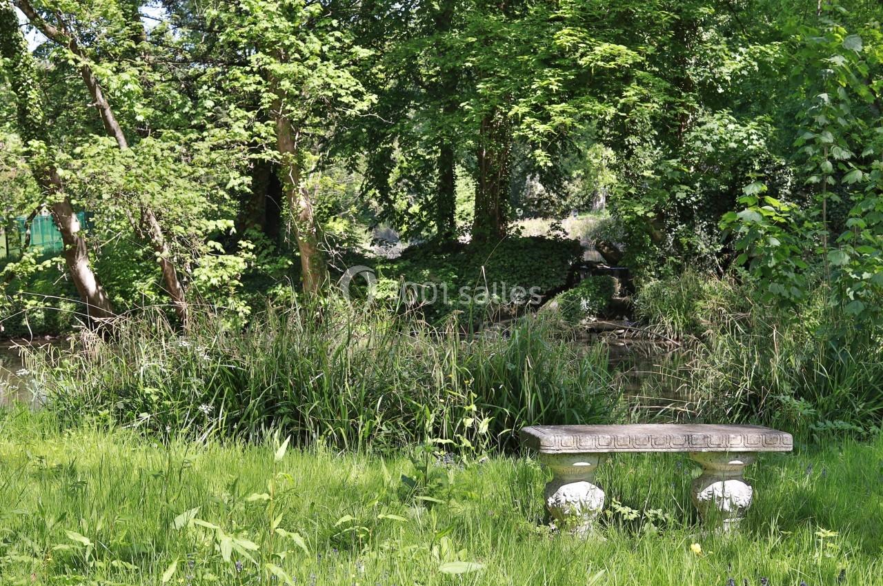 Banc en pierre dans un jardin verdoyant avec des arbres, des herbes hautes et un ruisseau en arrière-plan.