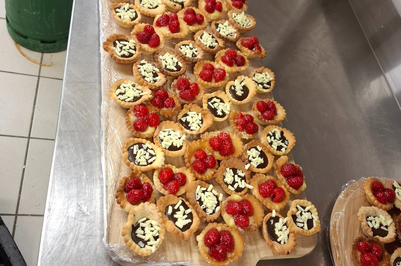 Plateau de tartelettes garnies de chocolat, éclats de chocolat blanc et framboises, posé sur un plan de travail en inox.