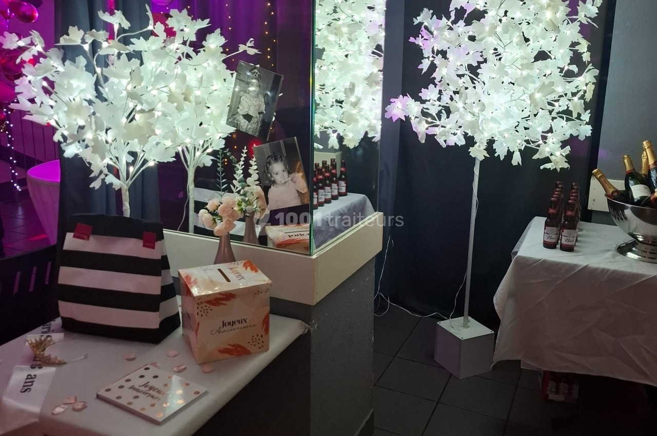 Décoration festive avec arbres lumineux blancs, photo encadrée, boissons et cadeaux sur une table.