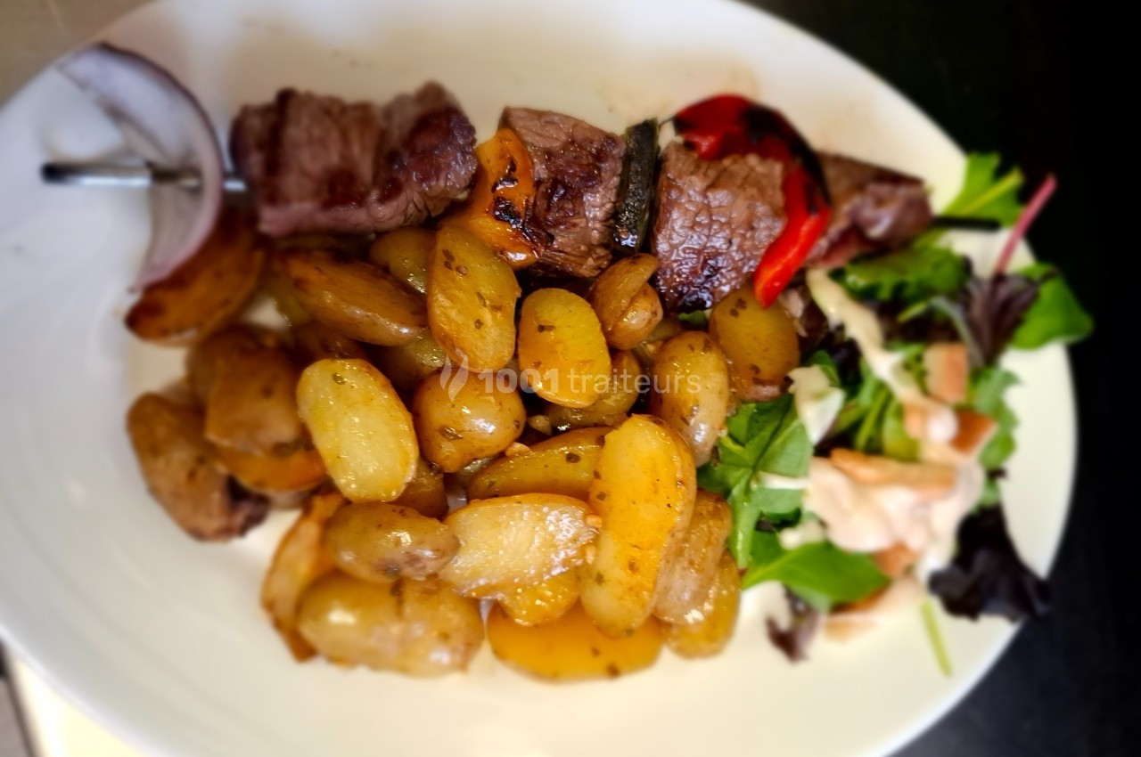Assiette contenant des pommes de terre rôties, morceaux de viande grillée, légumes et salade verte avec sauce.