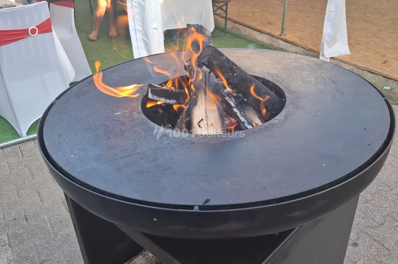 Brasero rond en métal noir avec des bûches enflammées, installé en extérieur sur un sol pavé.