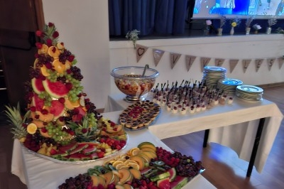 Buffet avec une pyramide de fruits frais, une salade de fruits, des verrines, des tartelettes et des assiettes empilées.