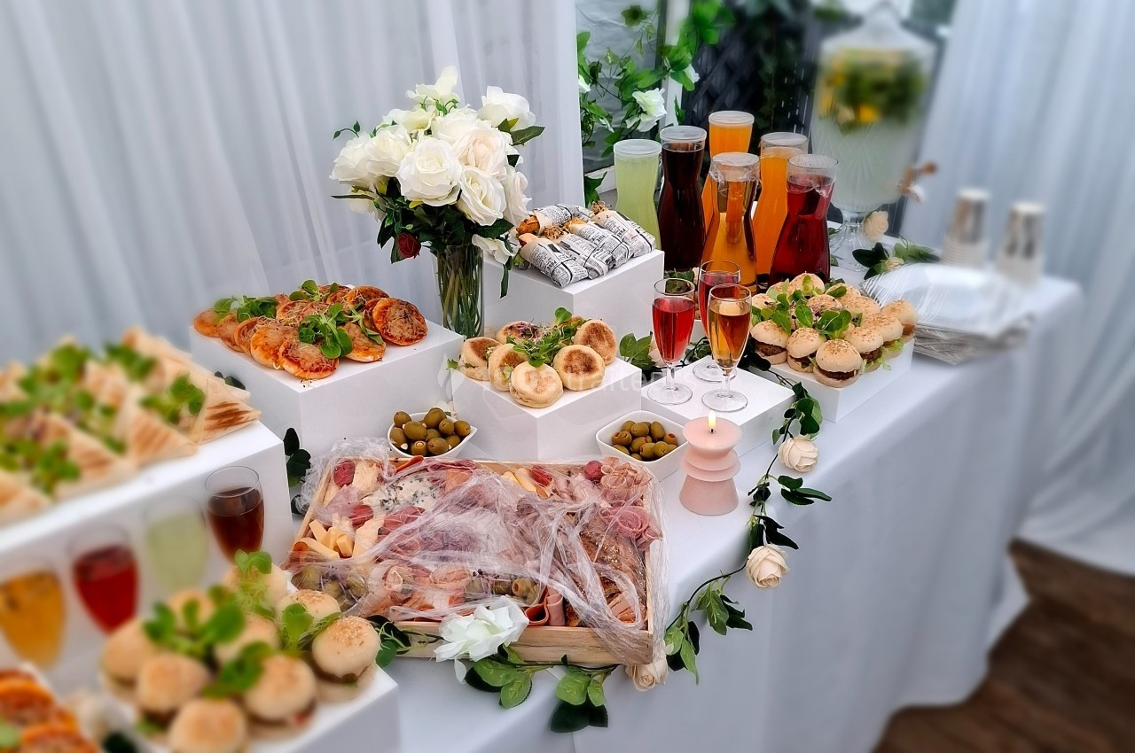 Buffet élégant avec assortiments de charcuterie, pains, olives, boissons variées et décorations florales blanches.