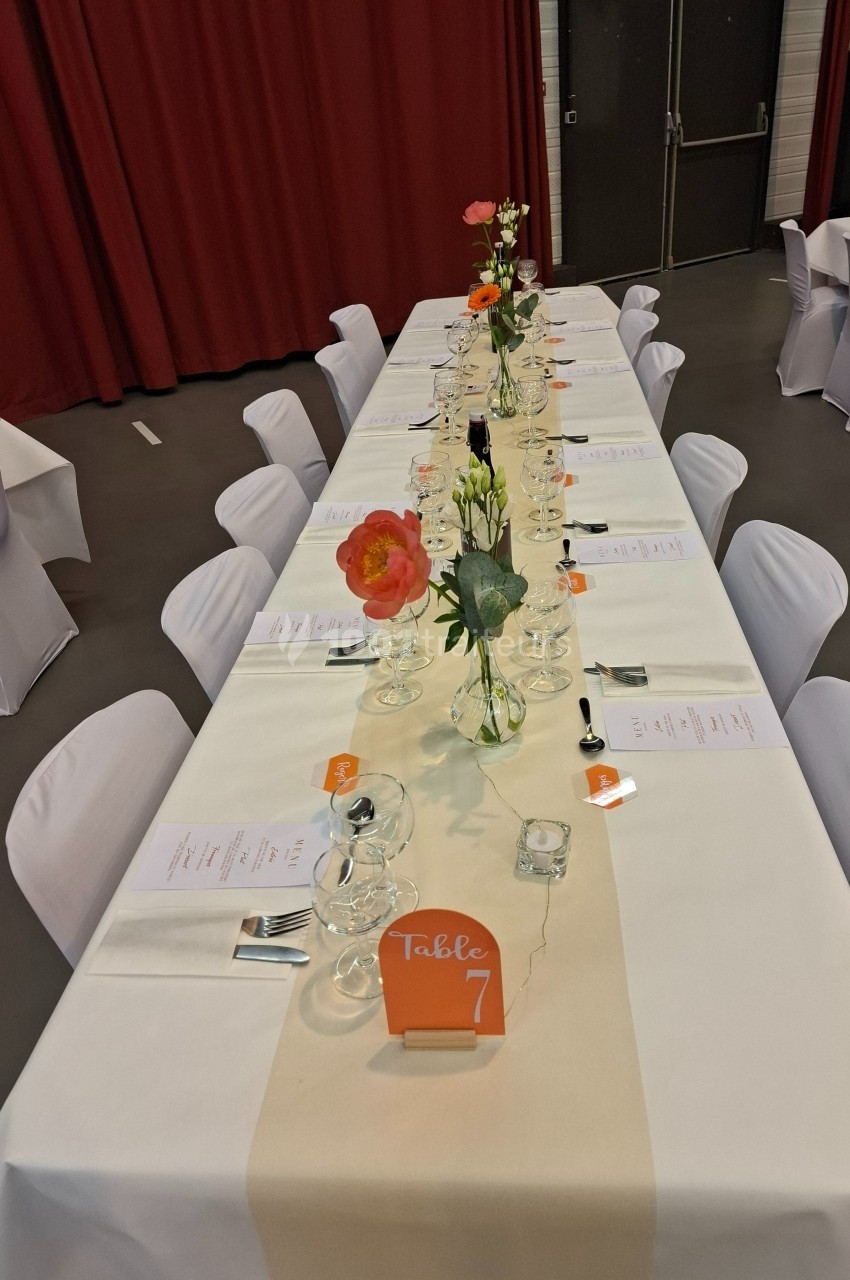 Table dressée avec nappes blanches, fleurs colorées, menus et numéro de table 7, dans une salle aux rideaux rouges.
