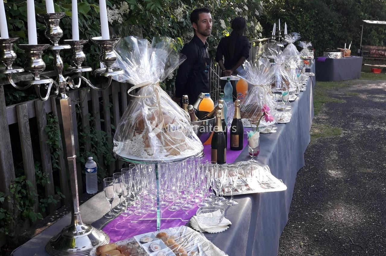 Buffet en extérieur avec boissons, viennoiseries, chandeliers et serveurs, disposé sur une longue table.