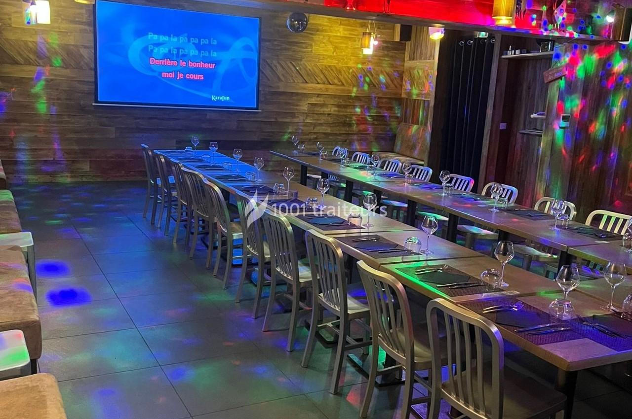 Salle de karaoké avec tables dressées, chaises alignées et éclairage coloré diffusé sur les murs et le sol.