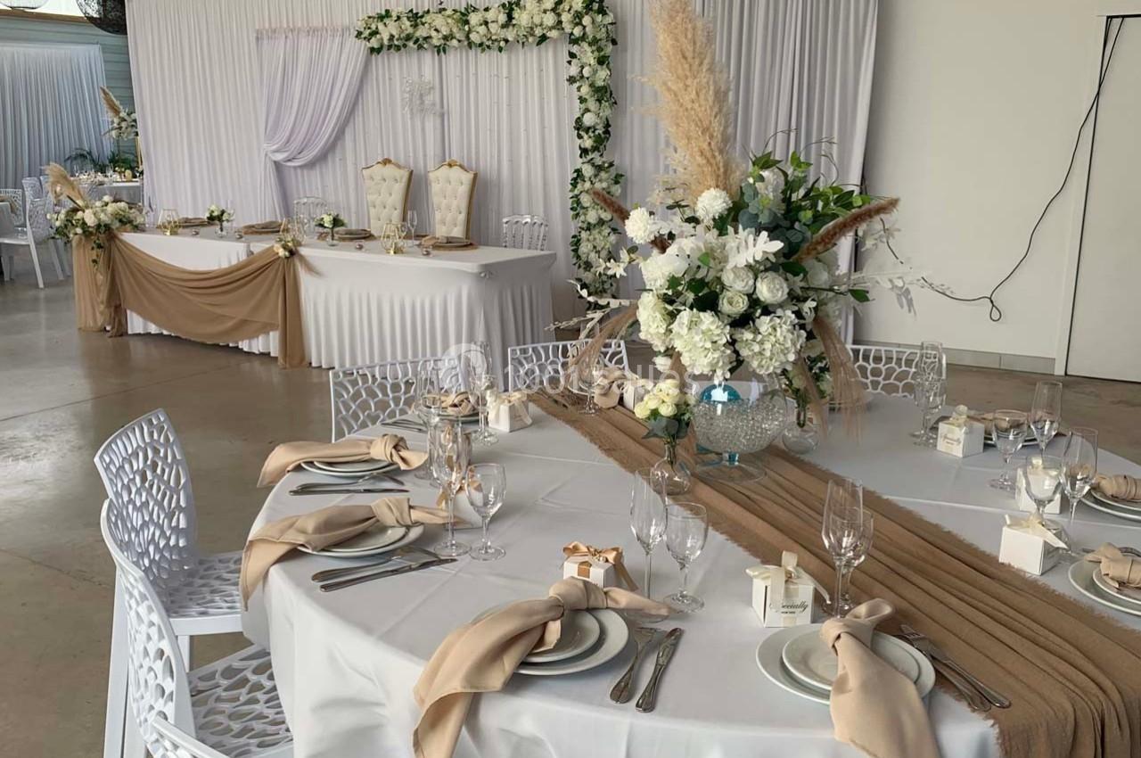 Salle de réception décorée pour un mariage avec tables dressées, nappes blanches, chemins de table beige et fleurs blanches.