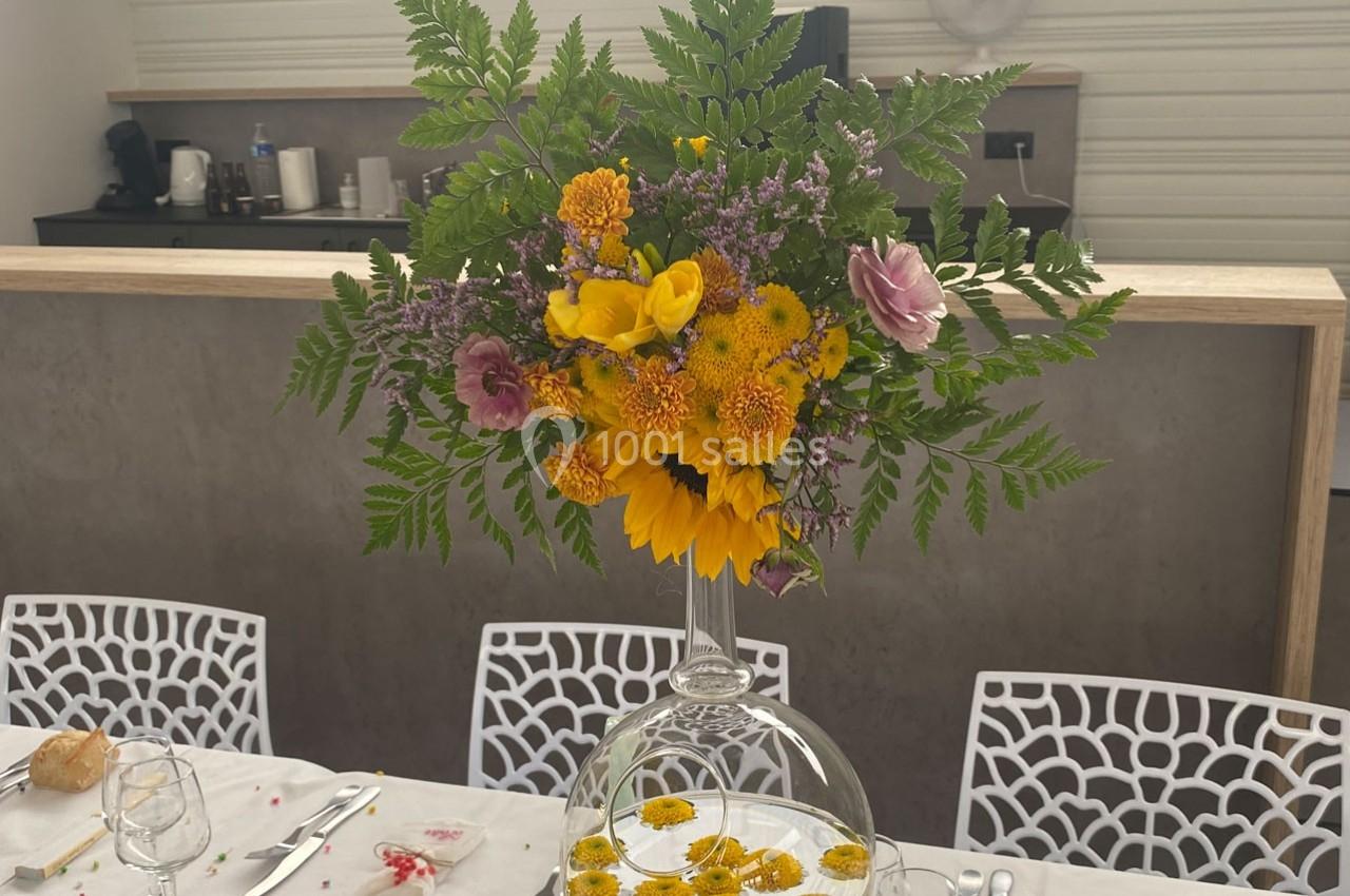 Centre de table floral avec tournesols, fougères et fleurs colorées, posé sur une table dressée dans une salle lumineuse.