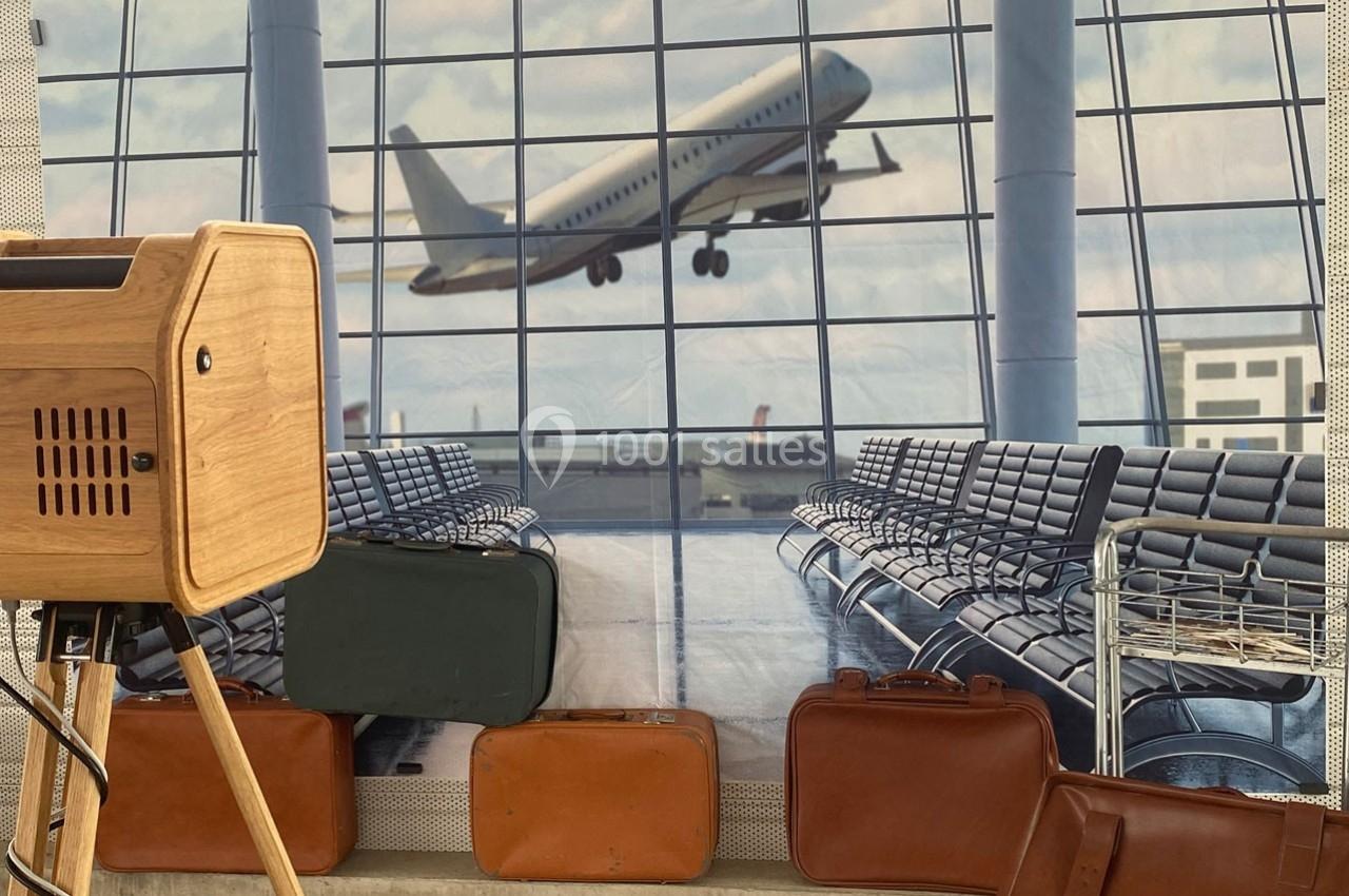 Un avion décolle vu à travers une grande baie vitrée d'aéroport, avec des valises et des sièges au premier plan.