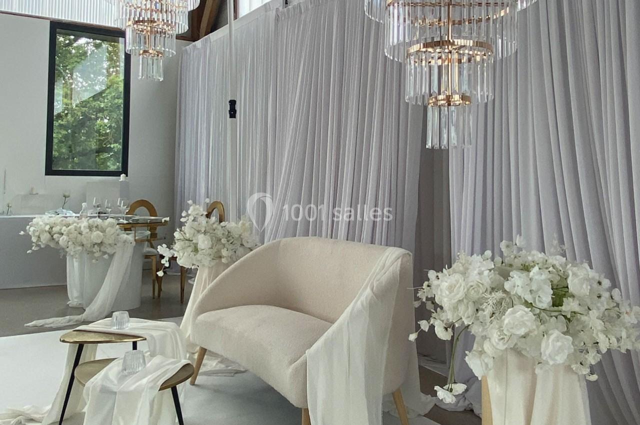 Salon décoré pour un événement, avec un canapé beige, des rideaux blancs, des fleurs blanches et des lustres élégants.