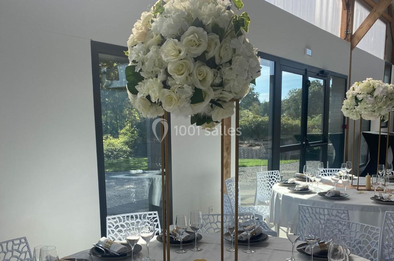 Centre de table floral avec roses blanches sur un support doré, dans une salle lumineuse avec grandes baies vitrées.