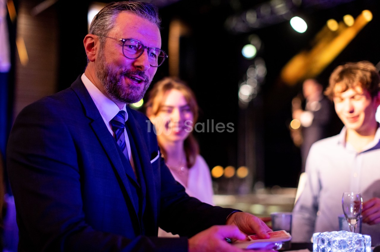 Un homme en costume effectue un tour de magie avec des cartes devant un petit groupe de personnes souriantes.