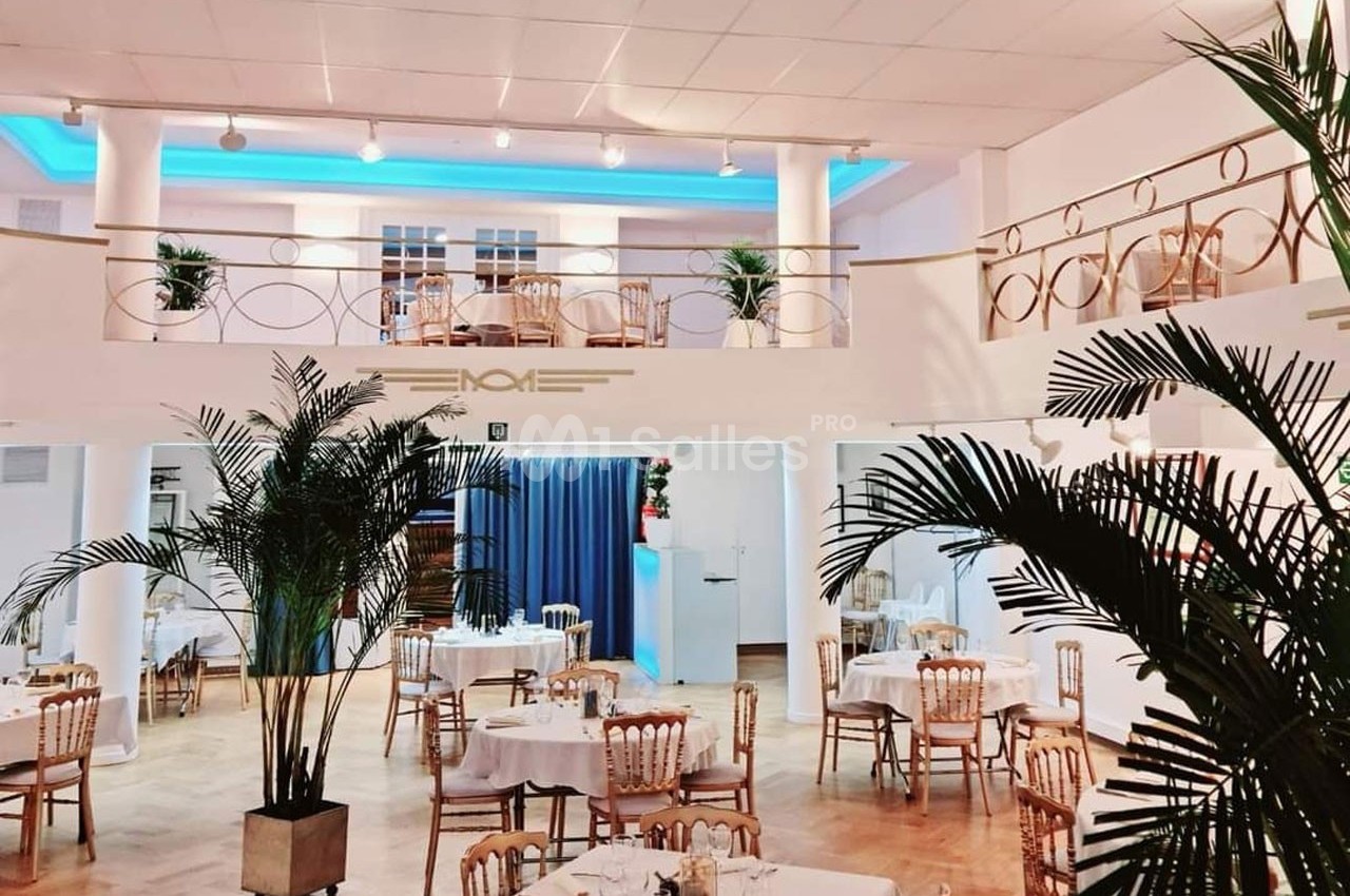 Salle de restaurant lumineuse avec tables dressées, plantes décoratives et mezzanine surplombant l'espace principal.