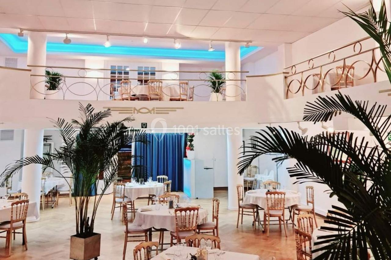 Salle lumineuse avec tables dressées, chaises en bois, plantes décoratives et mezzanine surplombant l'espace.