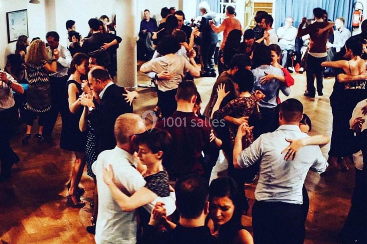 Des couples dansent dans une salle animée, éclairée par des lumières chaudes, sur un parquet en bois.