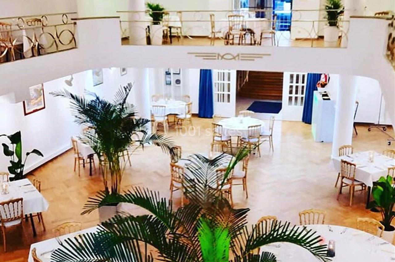 Salle lumineuse avec parquet, tables dressées et chaises dorées, décorée de plantes et dotée d'une mezzanine.