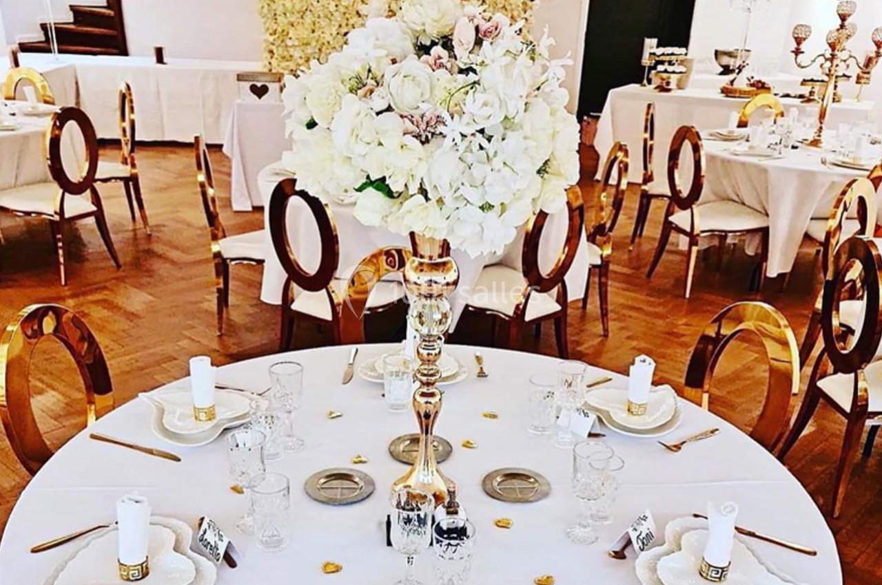 Salle de réception décorée avec des tables rondes, un centre de table floral et des chaises élégantes en bois doré.