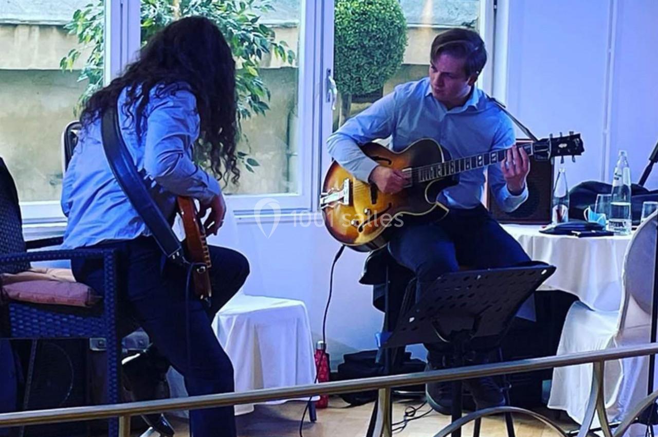 Deux musiciens jouent de la guitare dans une salle lumineuse, entourés de tables et de chaises.