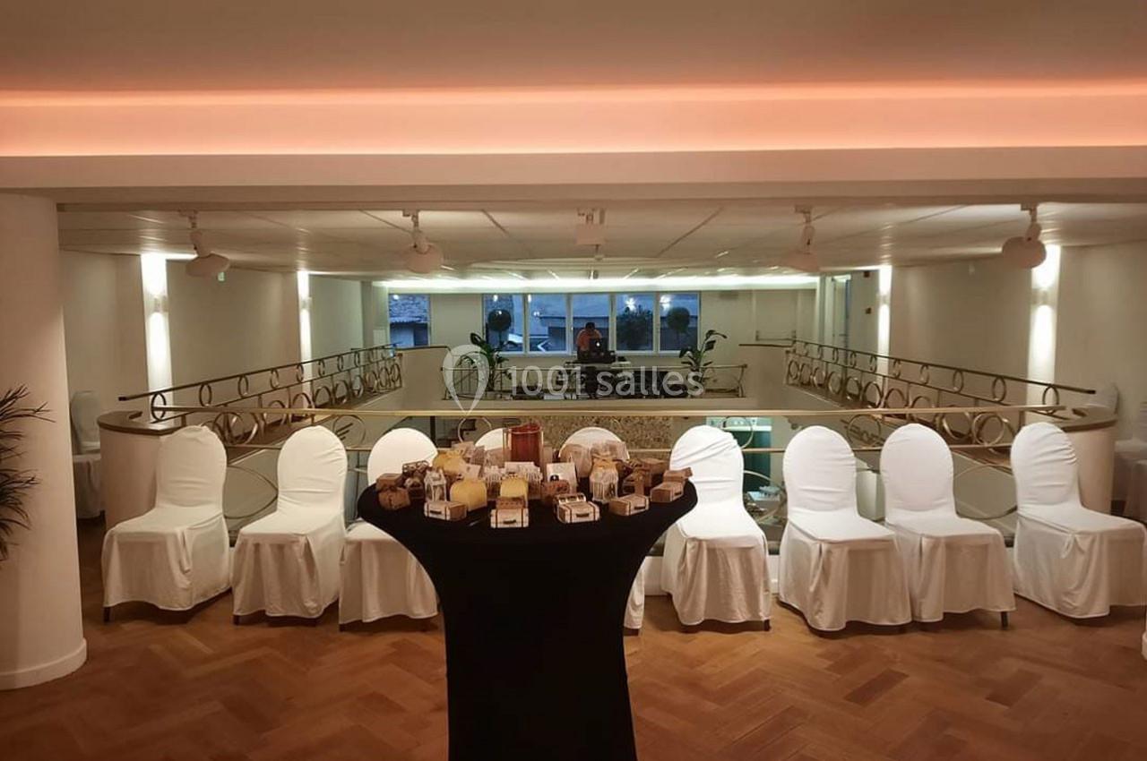 Salle lumineuse avec parquet, chaises blanches alignées et table haute noire garnie de petits objets ou collations.