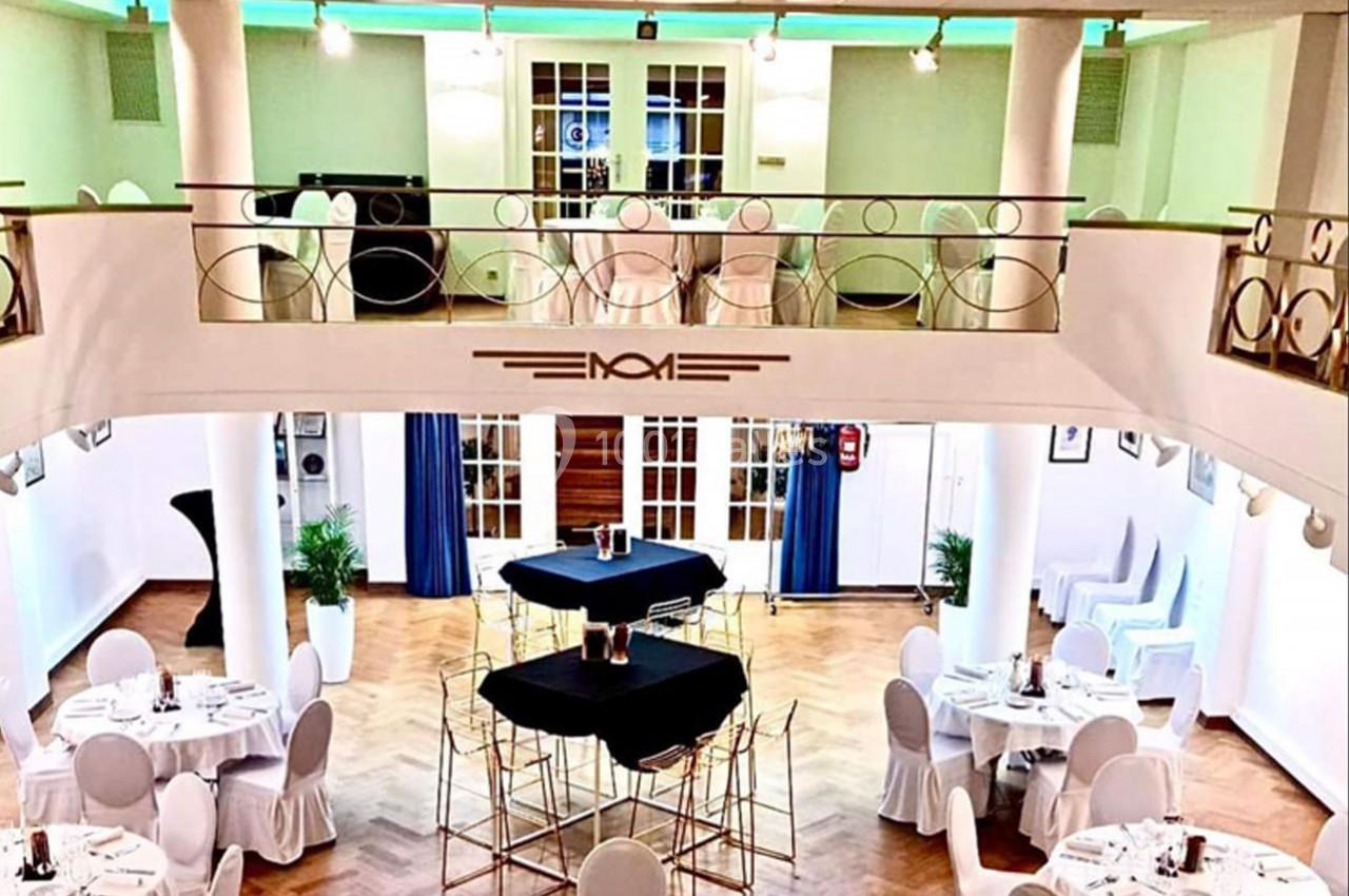 Salle de réception lumineuse à deux niveaux, avec tables rondes dressées et décorations élégantes.