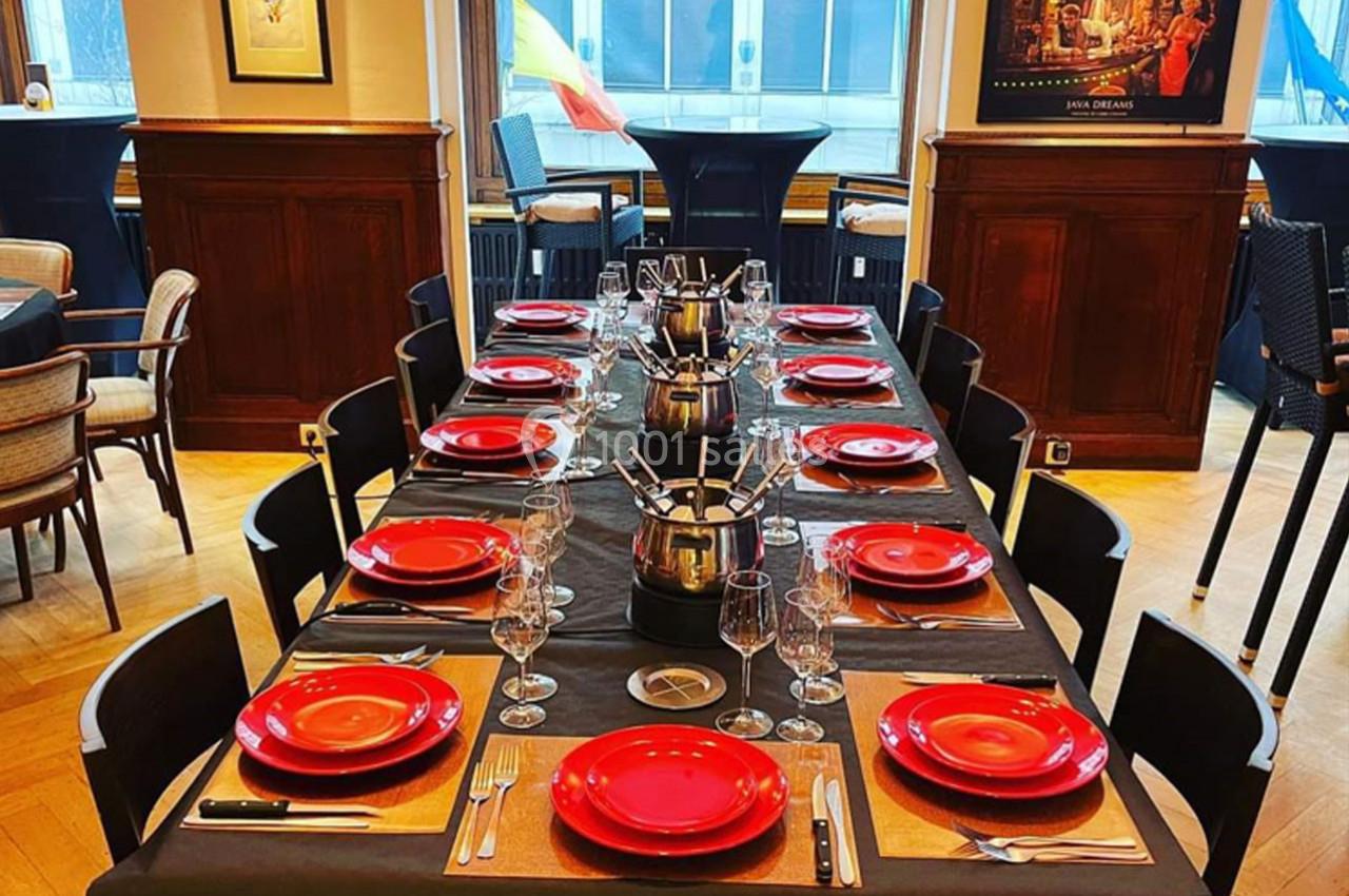 Table dressée avec des assiettes rouges, couverts et verres, dans une salle lumineuse au décor chaleureux.