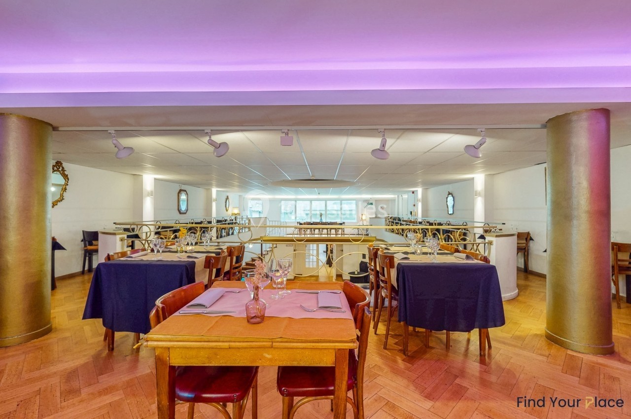 Salle de restaurant lumineuse avec tables dressées, chaises en bois et colonnes dorées, éclairage tamisé au plafond.