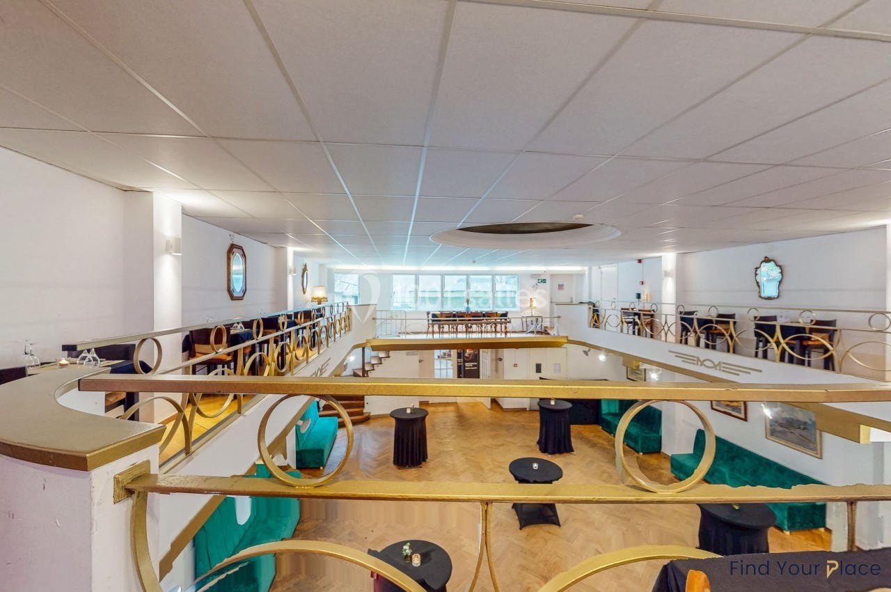 Salle lumineuse avec mezzanine, tables hautes noires, parquet en bois clair et décorations murales élégantes.