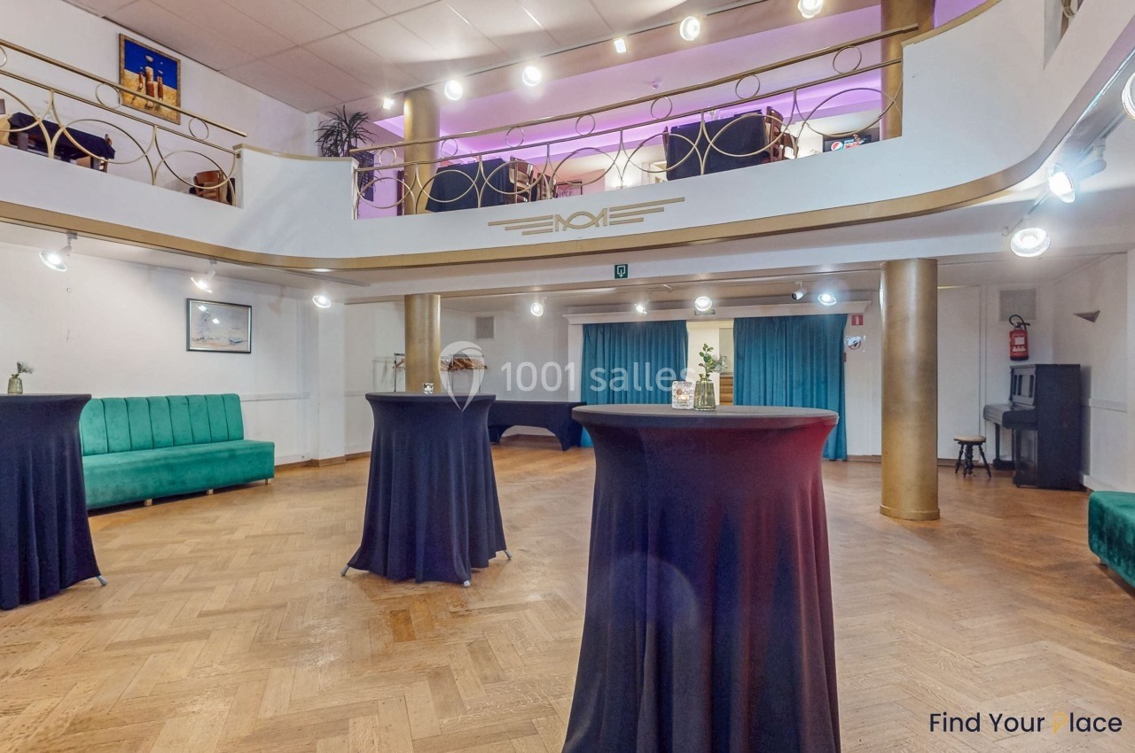 Salle lumineuse avec parquet, tables hautes drapées de noir, mezzanine et rideaux bleus en arrière-plan.