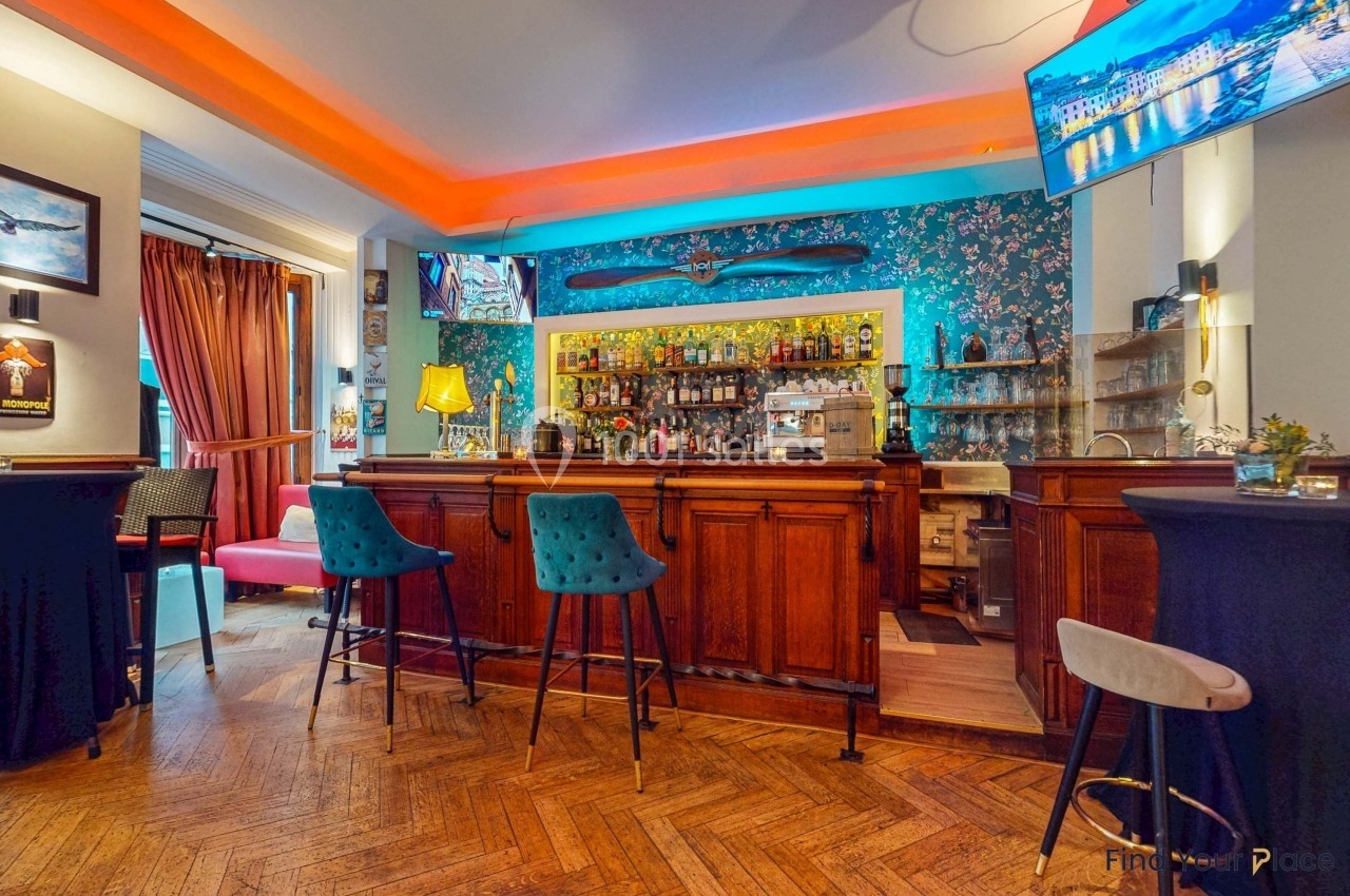 Bar avec comptoir en bois, tabourets colorés, étagères remplies de bouteilles et décor mural floral lumineux.