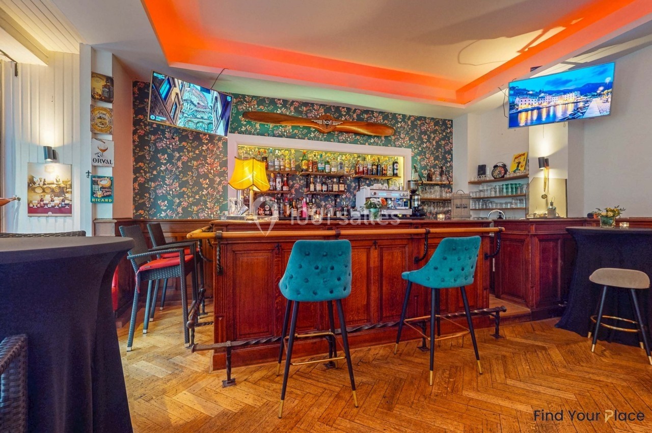 Bar avec comptoir en bois, tabourets colorés, étagères remplies de bouteilles et décoration murale florale.